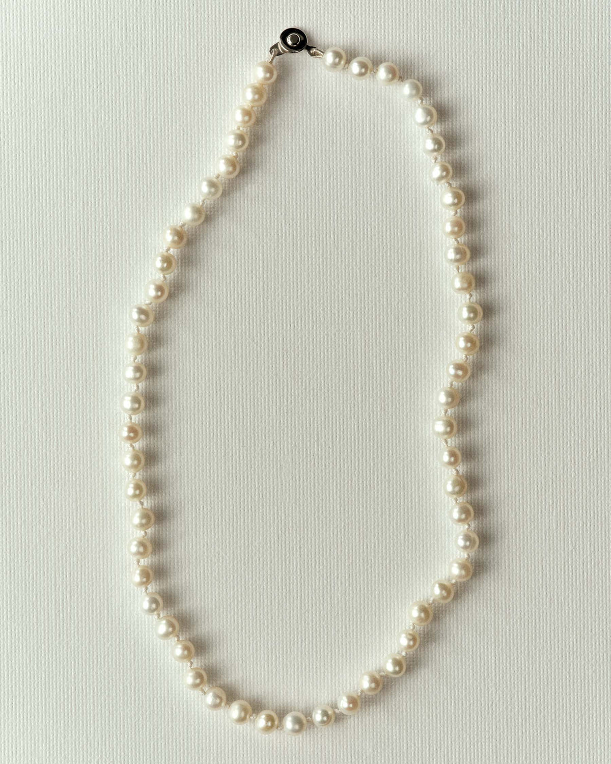 Strand of knotted cream pearls with a dark clasp, laid in an oval on off-white paper