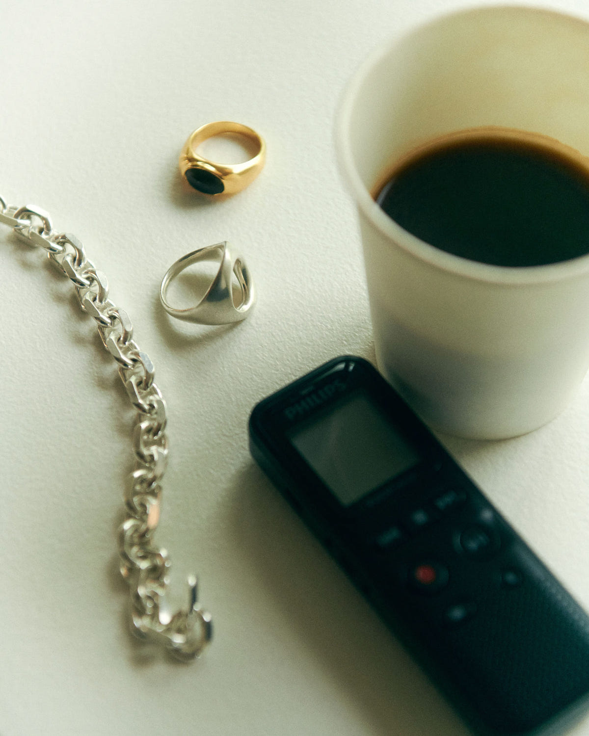 Chaîne argentée près d'une chevalière dorée à pierre noire, d'un café et d'un dictaphone