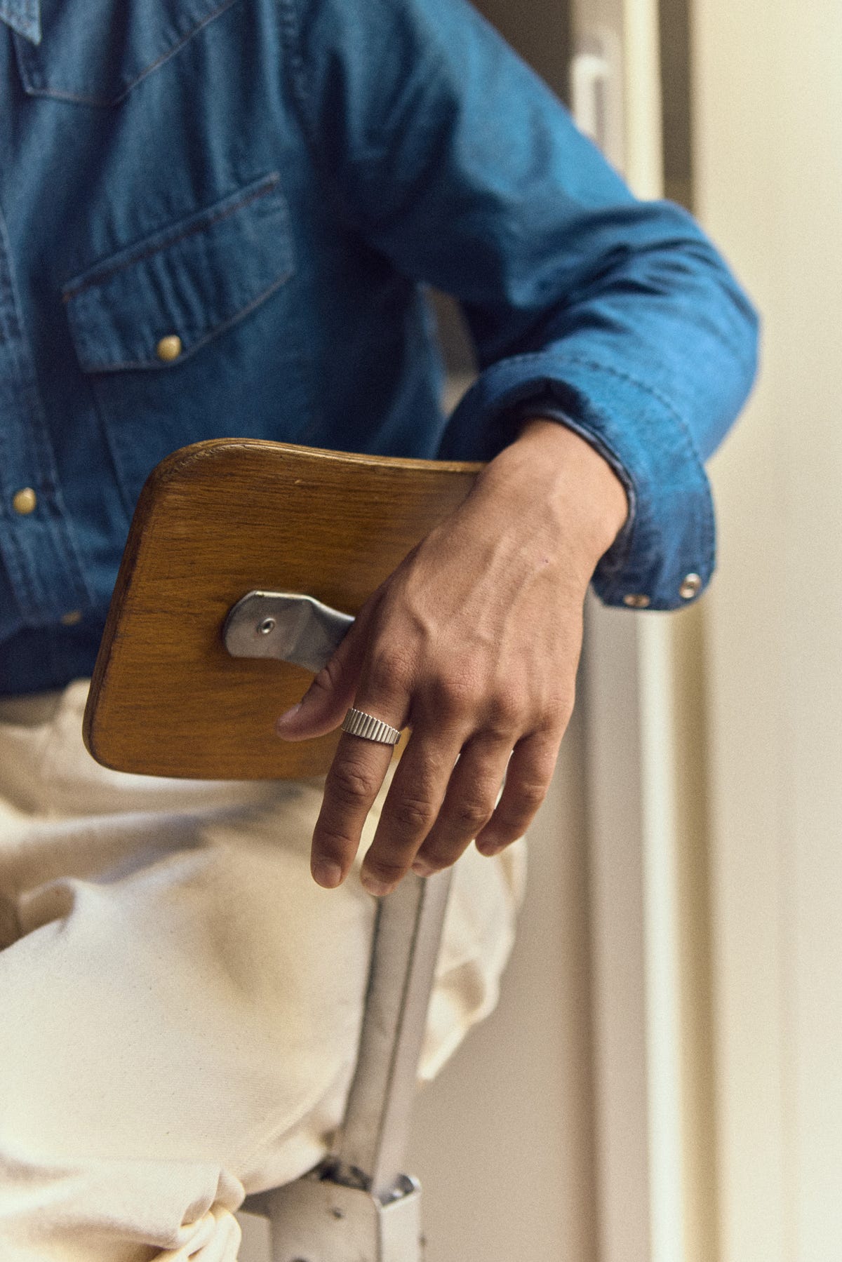 Silver ribbed band on the index finger, hand over a wood and metal chair back, denim shirt