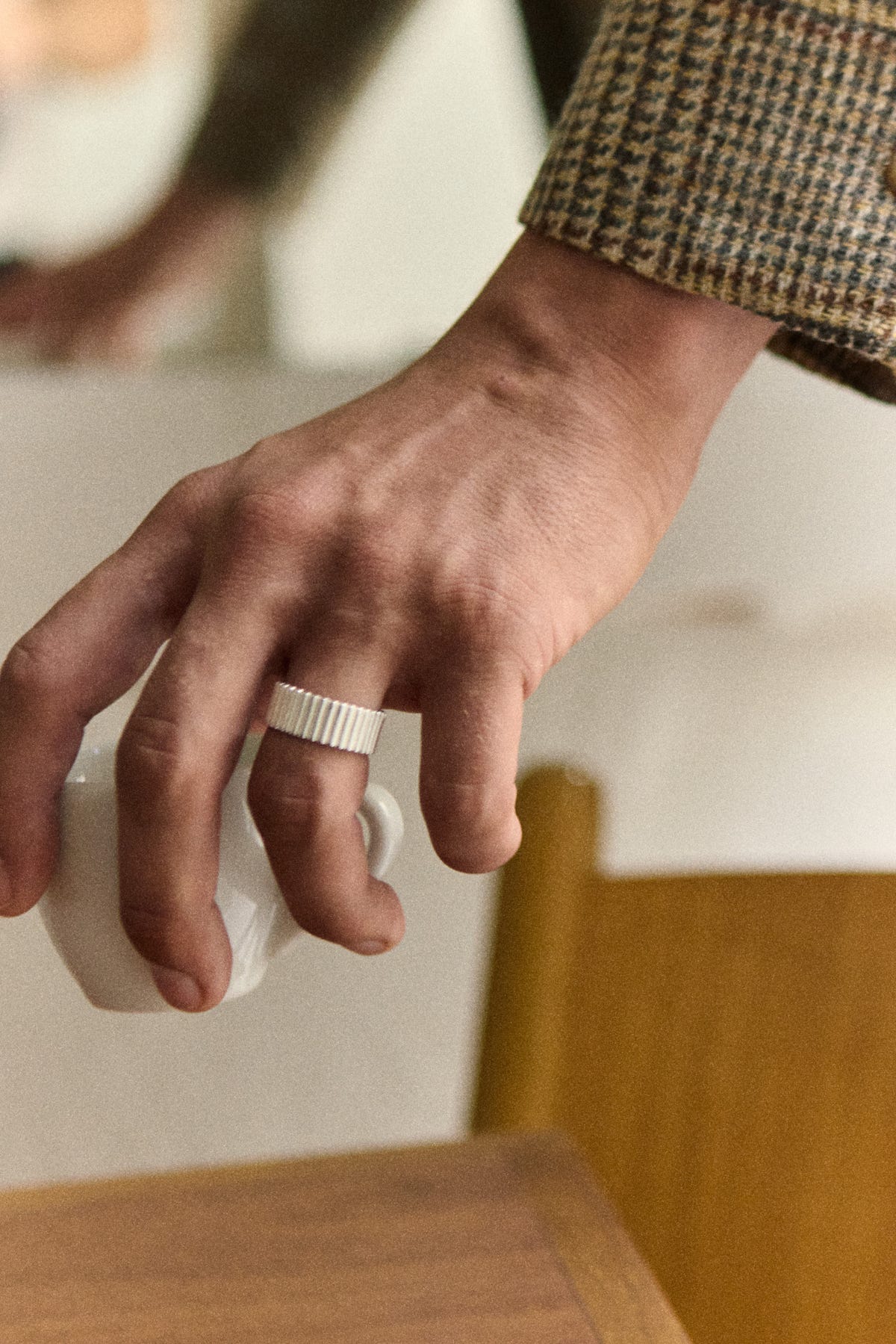 Wide silver ribbed band on a hand holding a white cup, check tweed sleeve, mustard chair