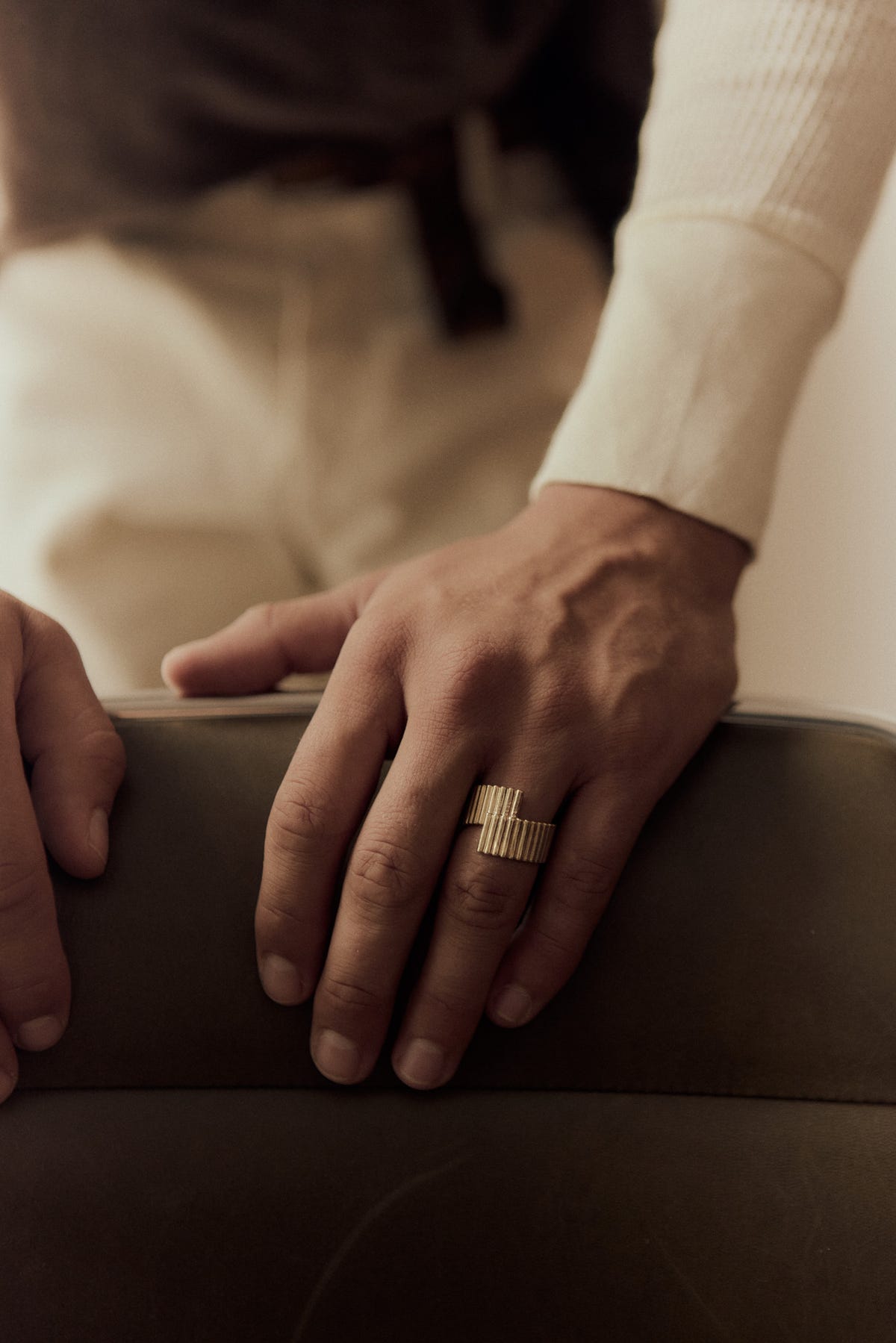 Gold ribbed ring with offset overlapping ends, both hands on a leather chair back, cream sleeve