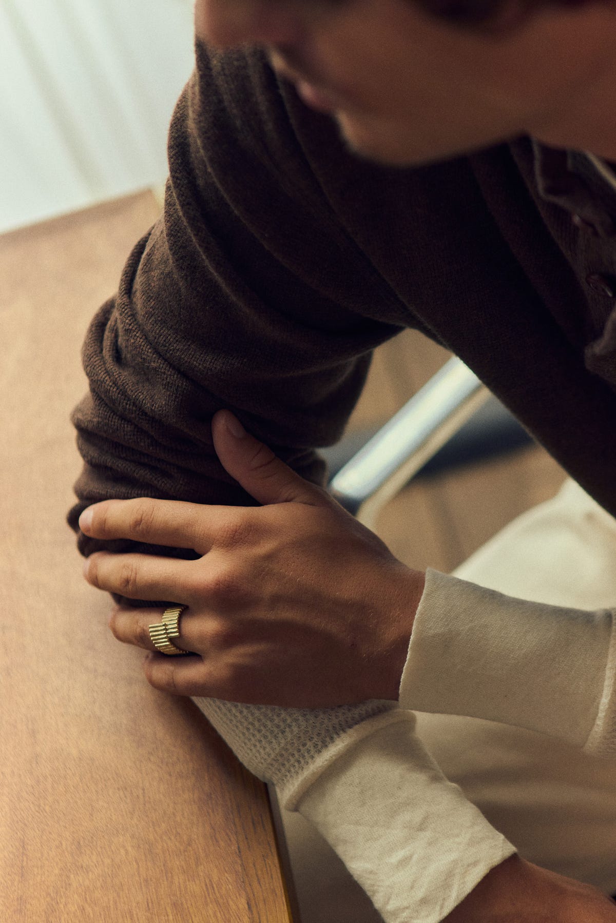 Gold ring worn, hand resting on a brown knit sleeve, arms on a wooden table