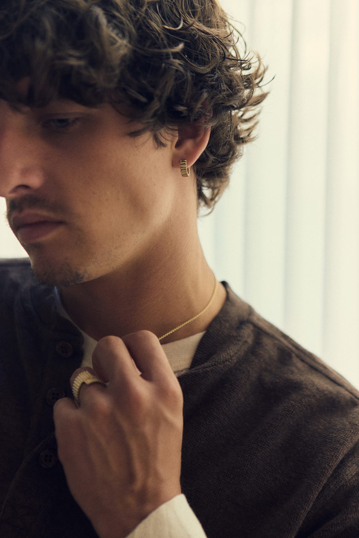 Gold hoop at the ear and ribbed gold ring on a hand holding a fine chain, brown henley
