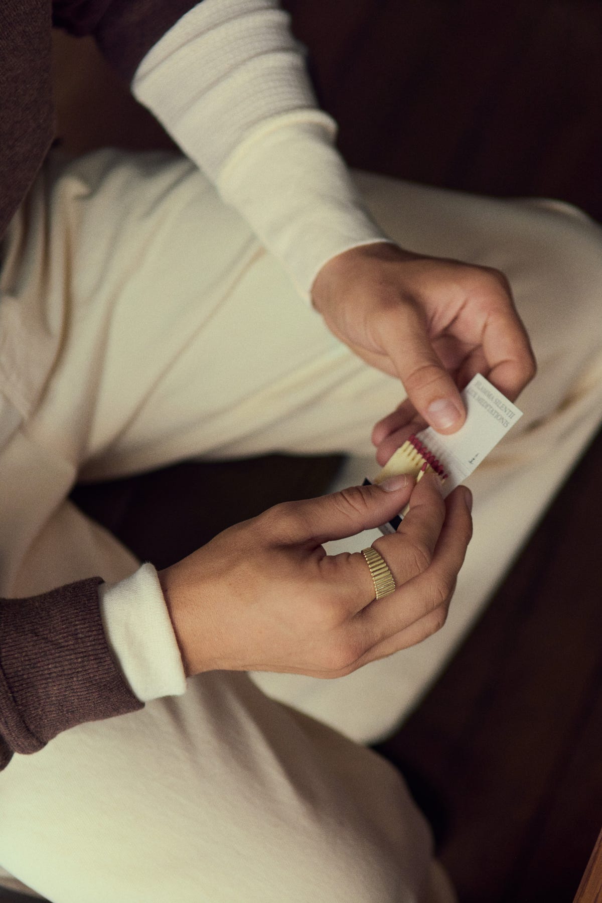 Gold ribbed band worn while opening a matchbook, cream sleeves and ecru trousers