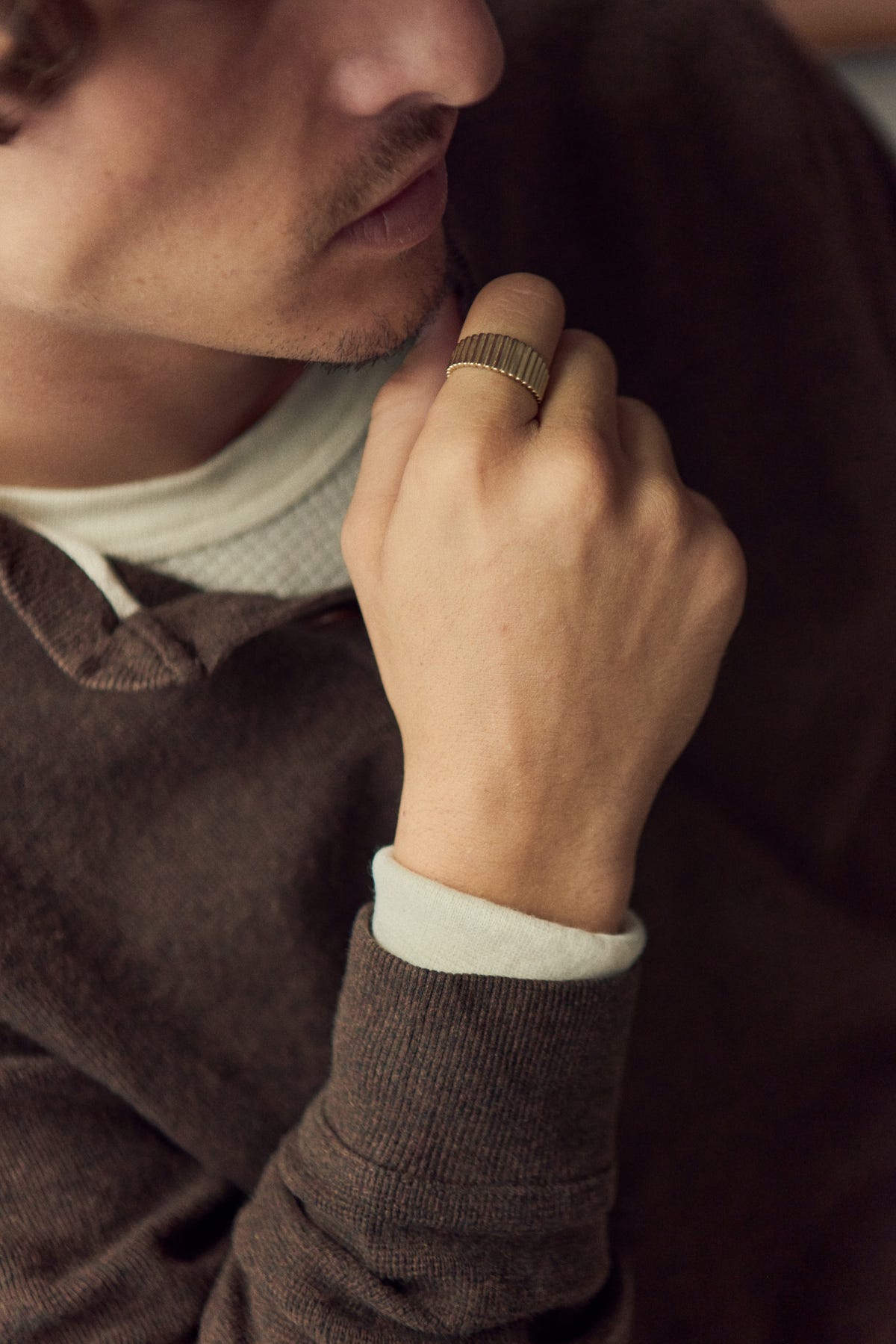 Gold ribbed band on the index finger, hand raised to the chin, brown knit over a cream top