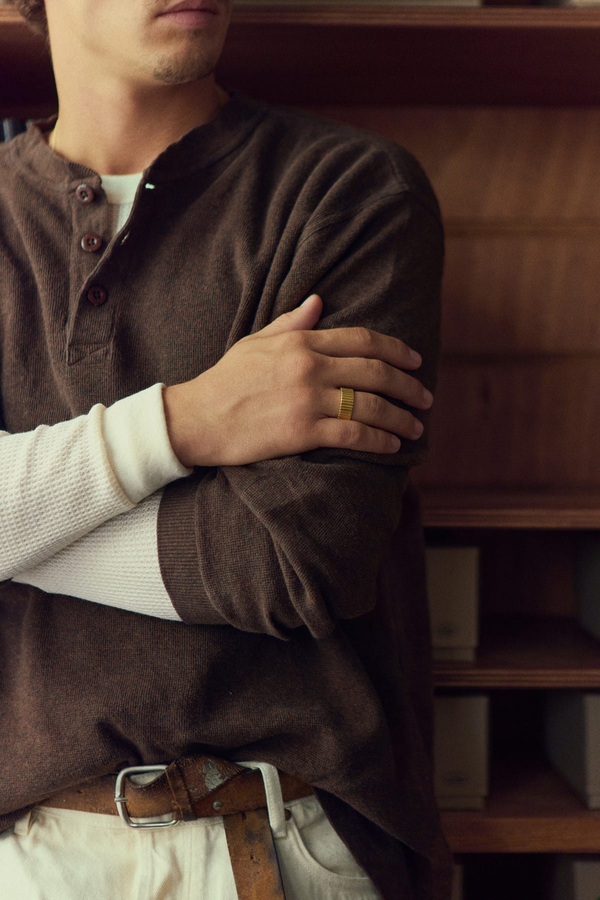 Wide gold ribbed band on a hand resting on a folded arm, brown henley, wooden shelves behind