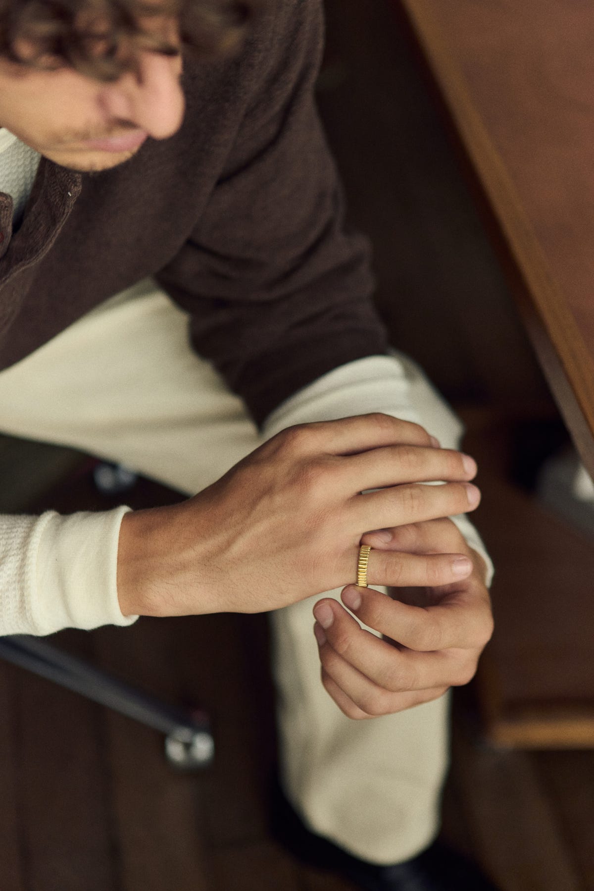 Gold ribbed band on clasped hands seen from above, brown sweater and ecru trousers