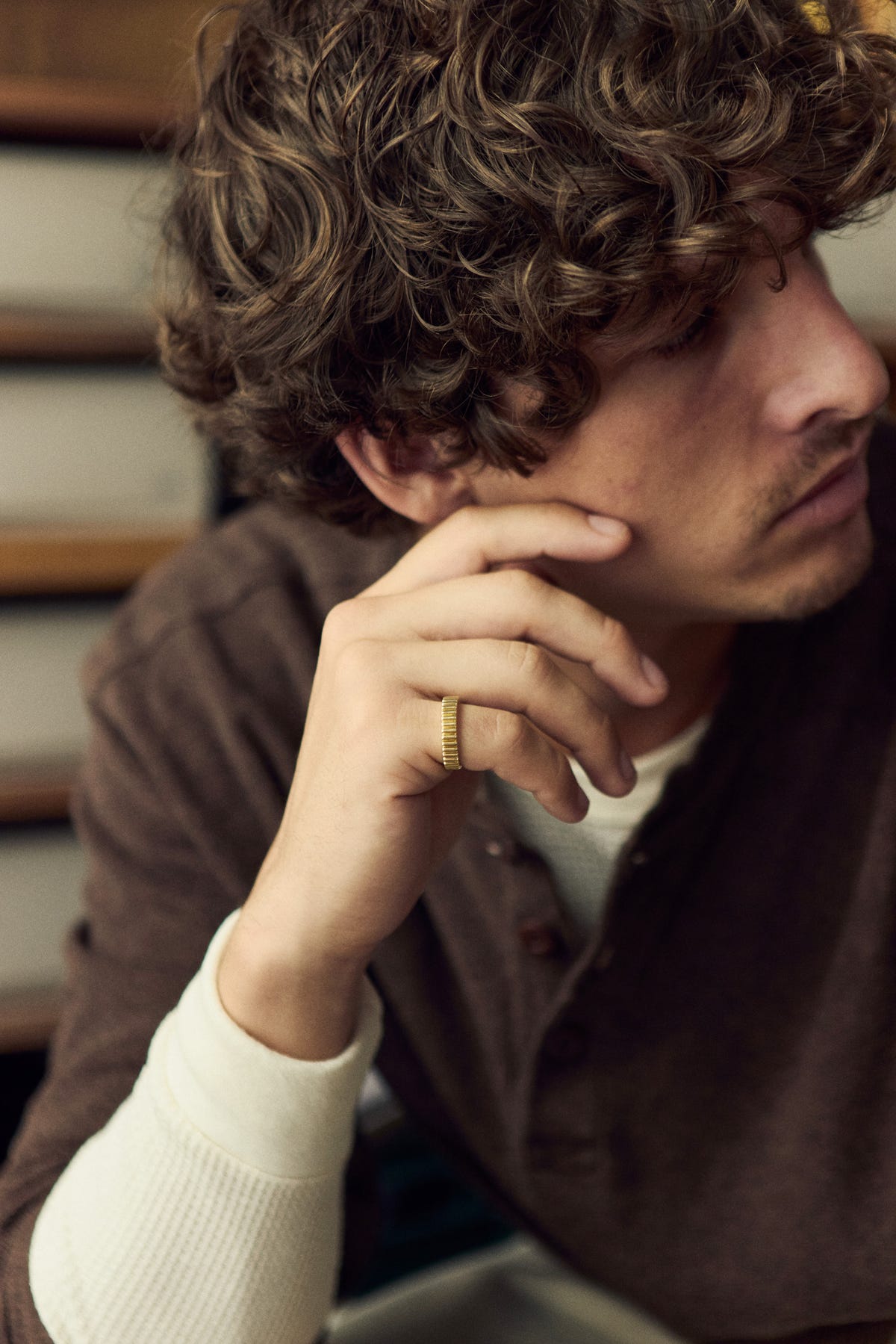 Gold ribbed band on a hand resting against the cheek, brown henley over a cream waffle top
