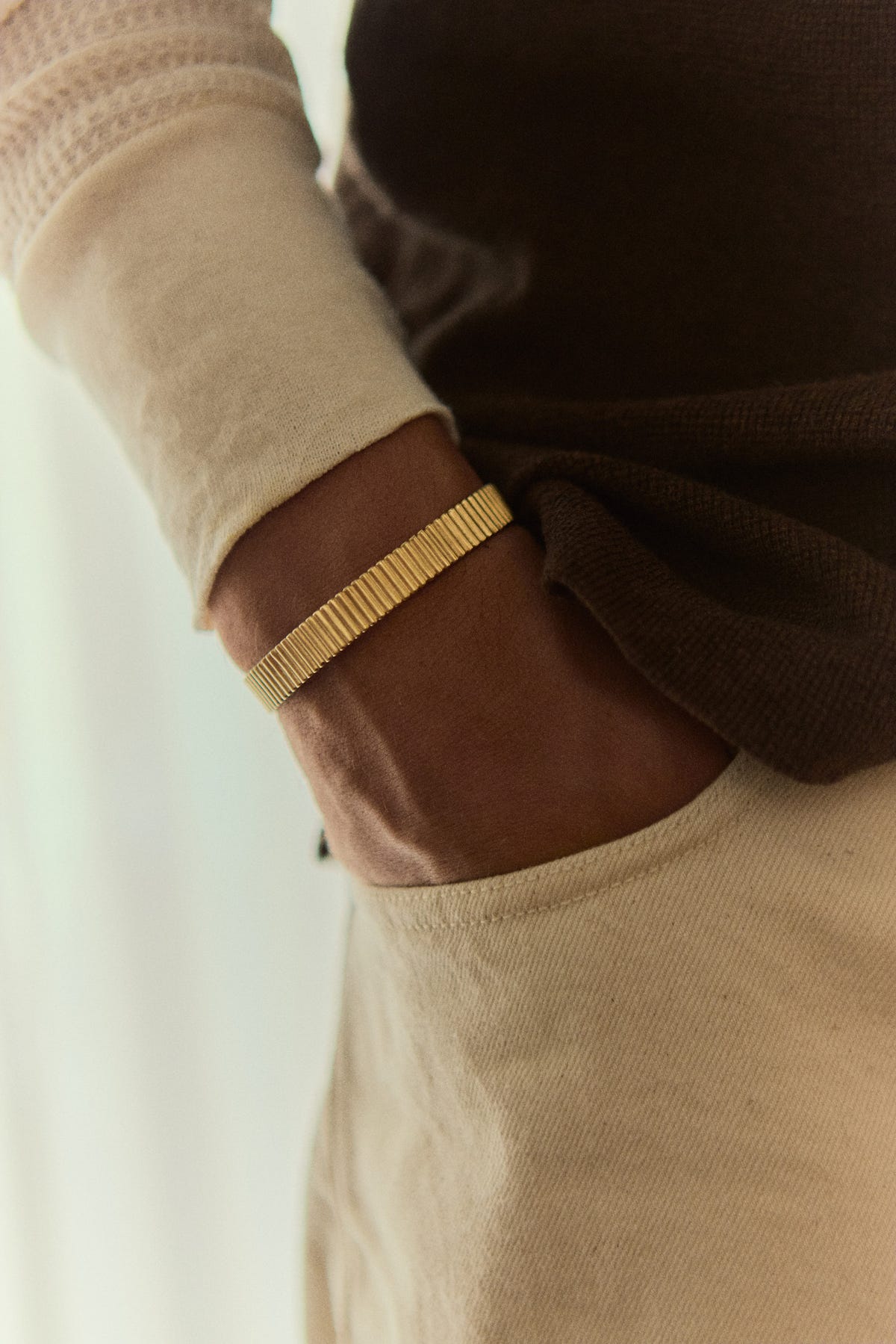 Wide ribbed gold cuff on the wrist, hand in a pocket, brown knit over a cream long sleeve