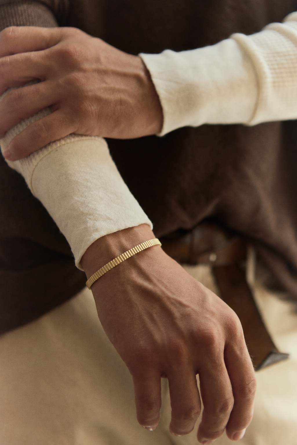 Thin ribbed gold cuff on the wrist, arms crossed, cream waffle-knit sleeves and brown sweater
