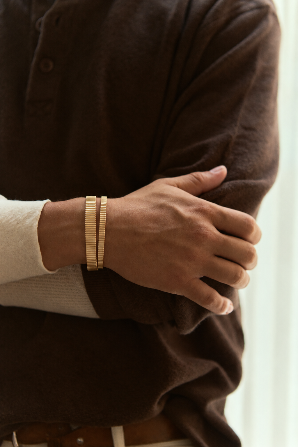 Two ribbed gold cuffs, one wide and one thin, on the same wrist, arms crossed, brown henley