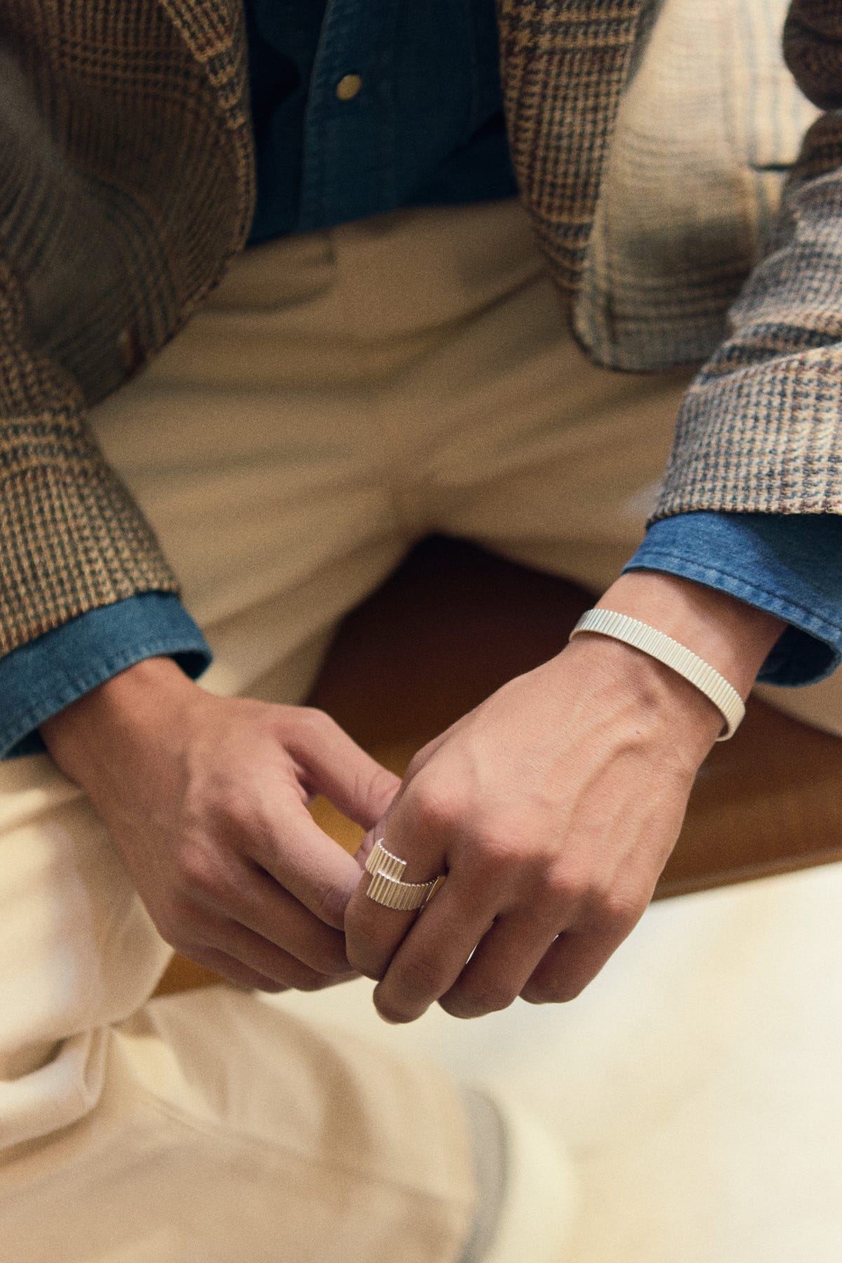 Ribbed silver cuff and ring worn on the same hand, check jacket over a denim shirt, seated