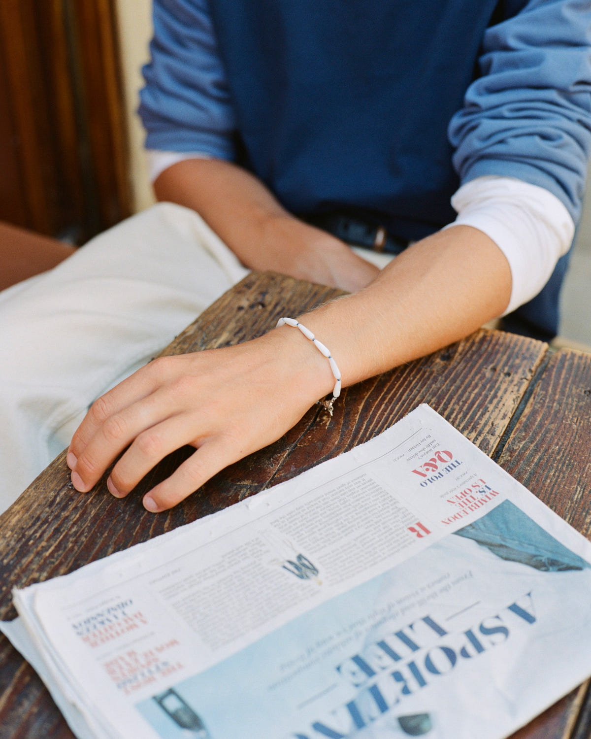 Mother-of-pearl drop bead bracelet on the wrist, hand resting on a newspaper on a wooden table
