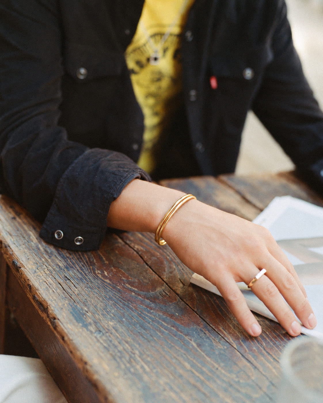 Gold-plated double cuff 3mm on the wrist with a gold signet ring, hand resting on a wooden table