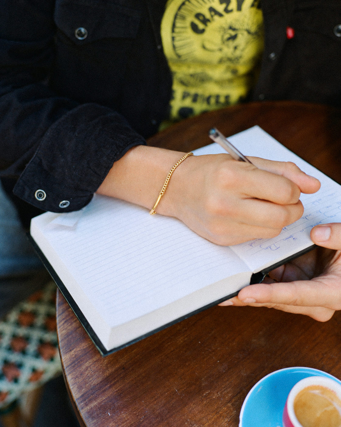 Gold-plated thin curb identity bracelet on the wrist, hand writing in a notebook at a cafe