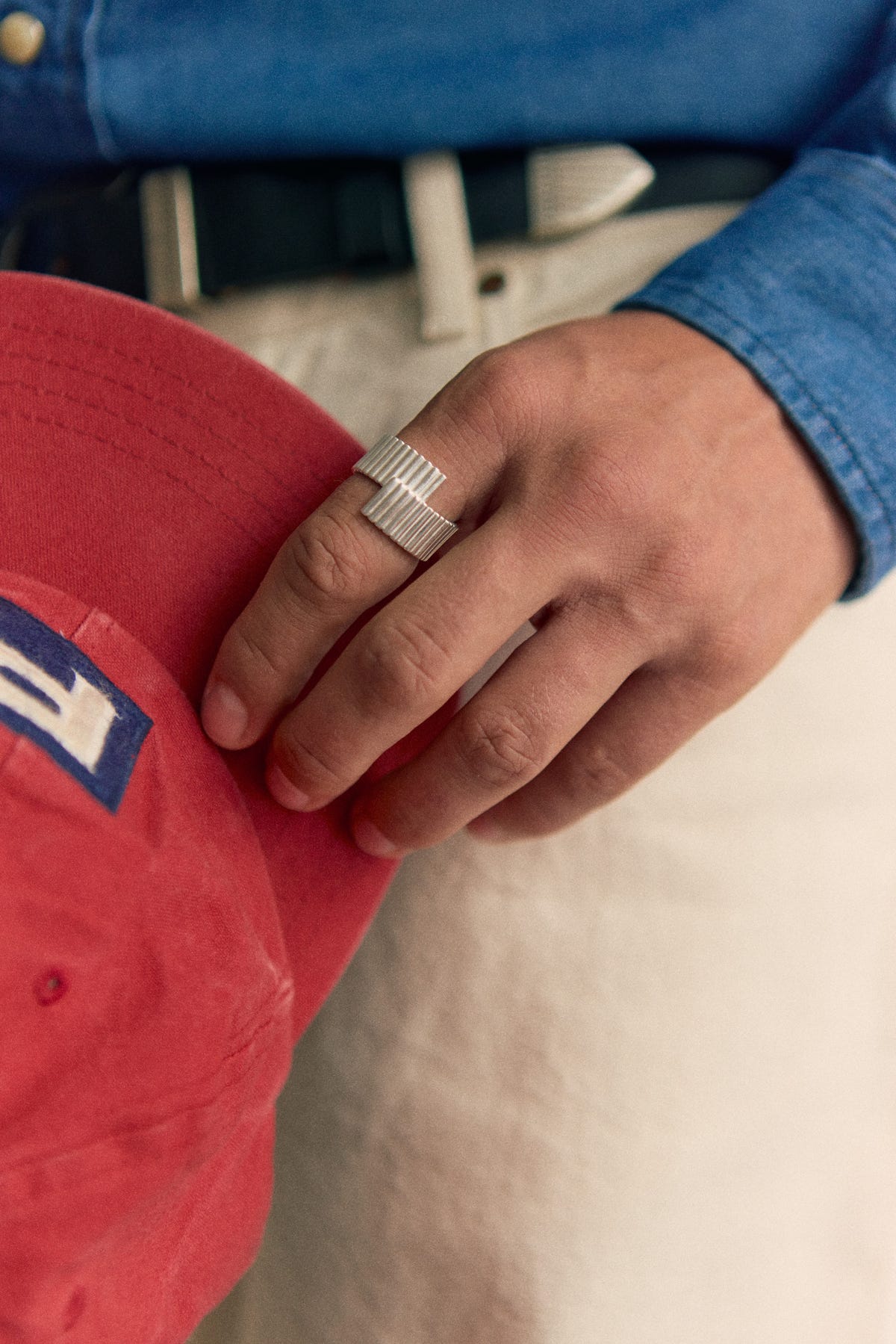 Silver ribbed ring with offset overlapping ends, hand holding a red cap, denim shirt