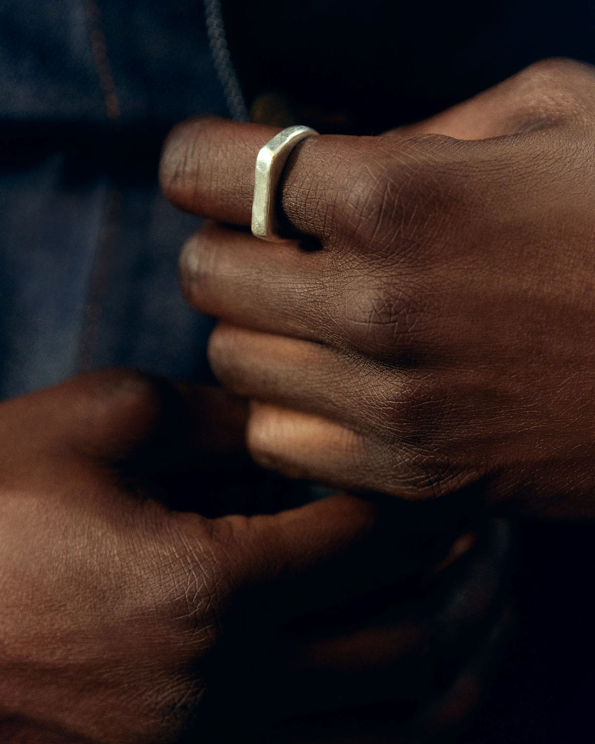 Silver signet worn on the finger, hands clasped against dark denim