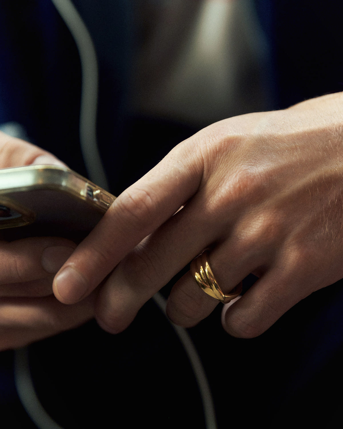 Gold band with two crossing strands worn on the ring finger, hands holding a phone