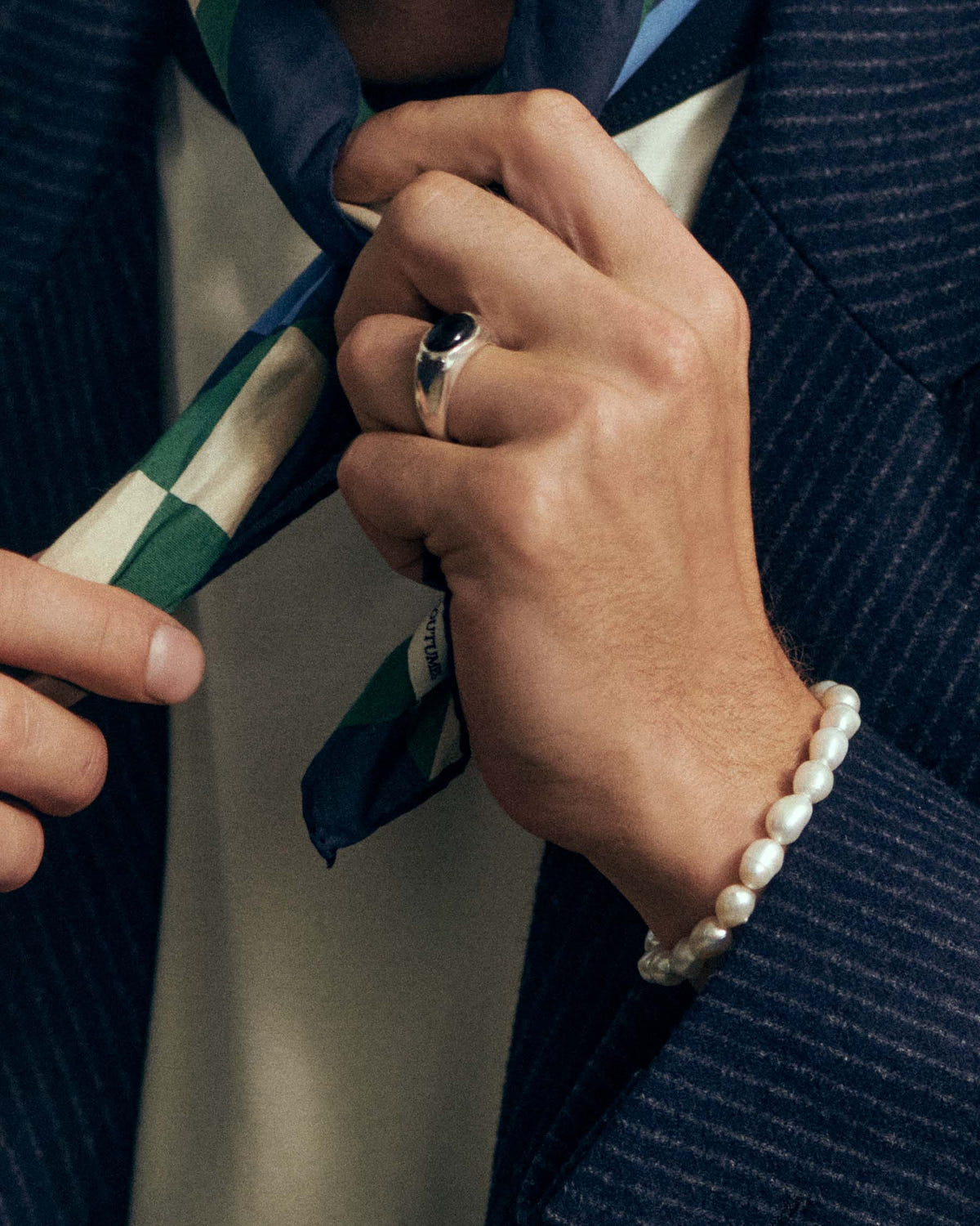 Perles crèmes au poignet, mains nouant un foulard à damiers sur veste marine rayée