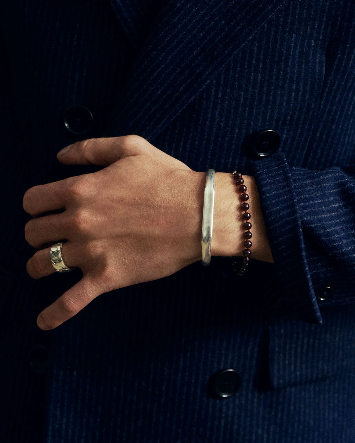 Wrist resting on a navy pinstripe jacket, dark beads, silver cuff and gold ring