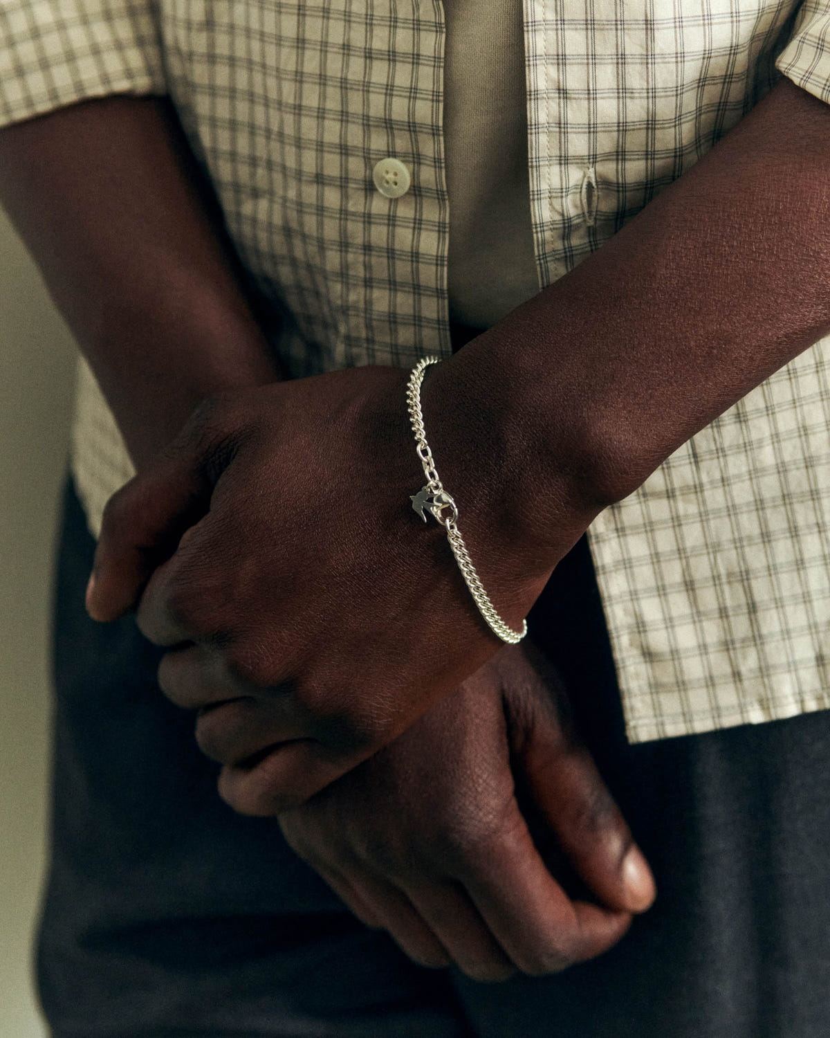 Silver curb links with a bird charm at the wrist, hands crossed, checked shirt