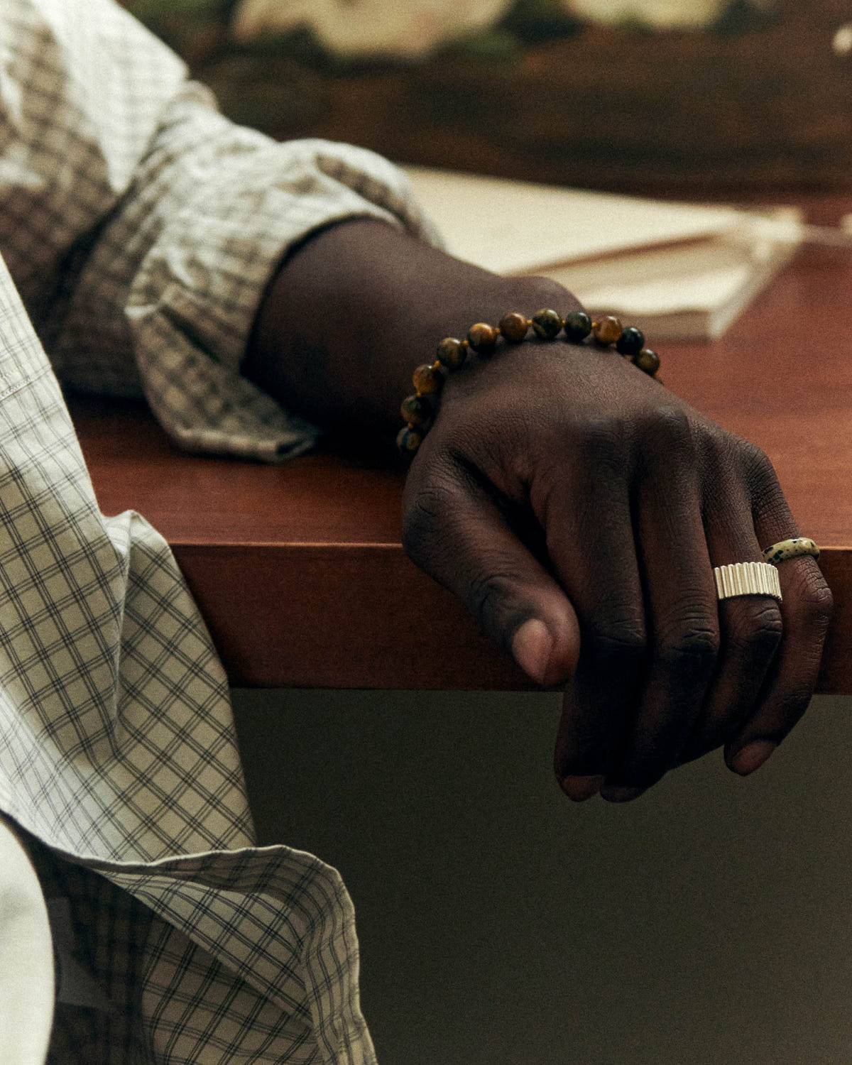 Wide silver ribbed band on a hand resting on a wooden desk, tiger's eye bead bracelet, check shirt