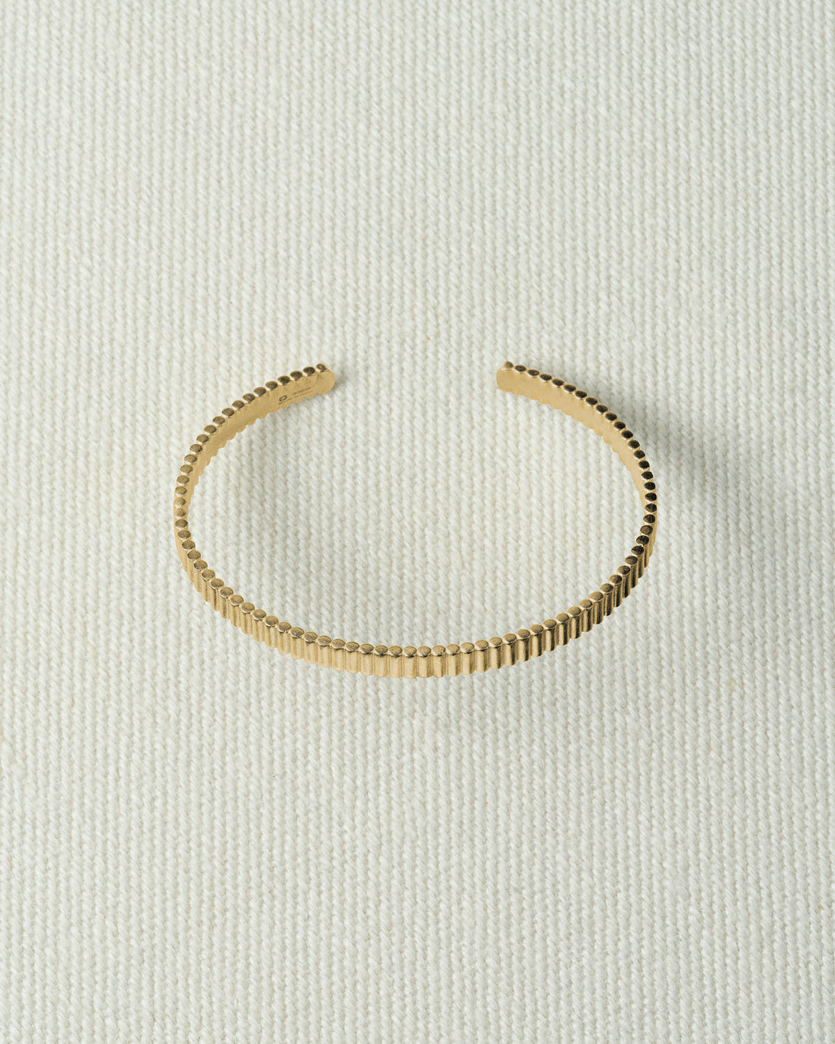 Thin ribbed gold cuff, open at the back, lying flat on textured ecru canvas