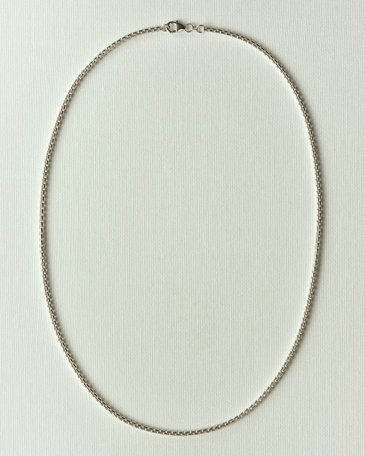 Silver box-link chain laid in an oval on off-white paper, clasp at the top