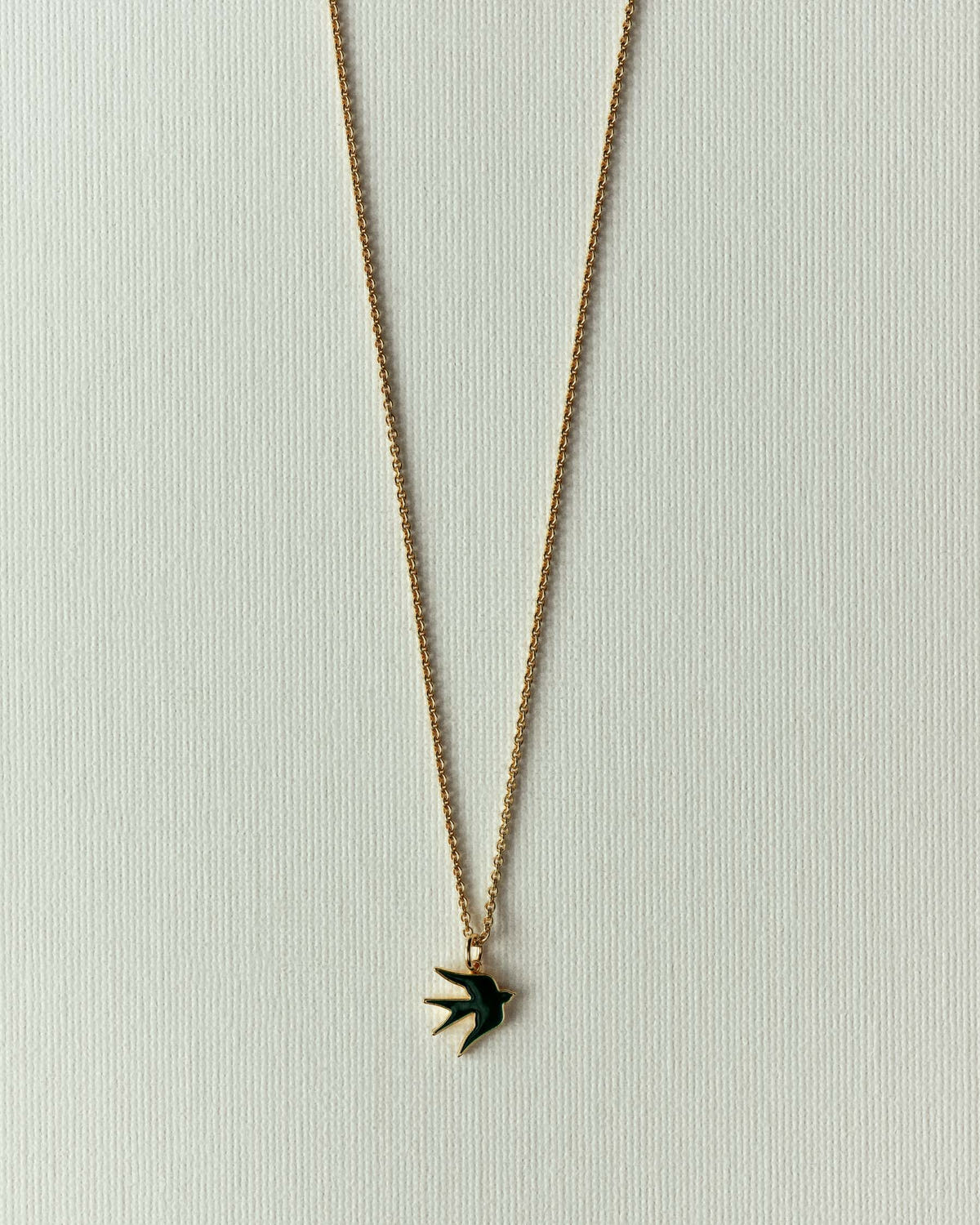 Fine gold chain with a small dark green enamelled bird, flat on off-white paper