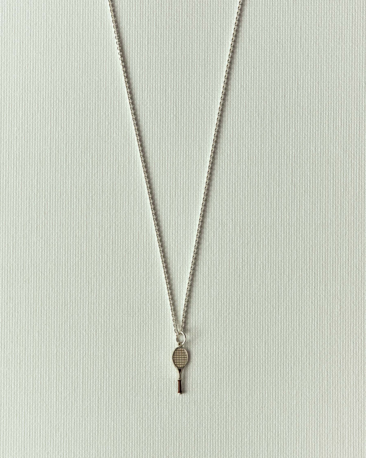 Tiny silver tennis racket charm on a fine chain, off-white paper background