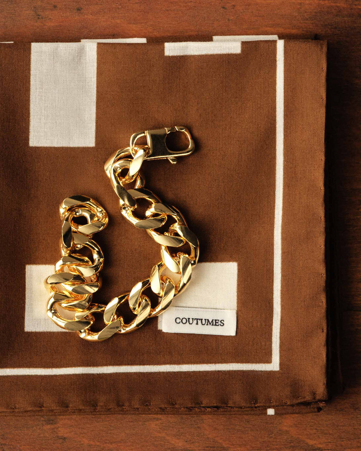 Brown square folded flat, chunky gold curb bracelet placed on top, Coutumes label