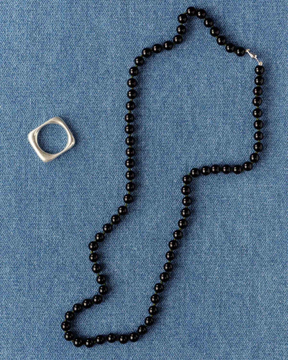 Glossy black beads coiled on blue denim beside a square silver ring